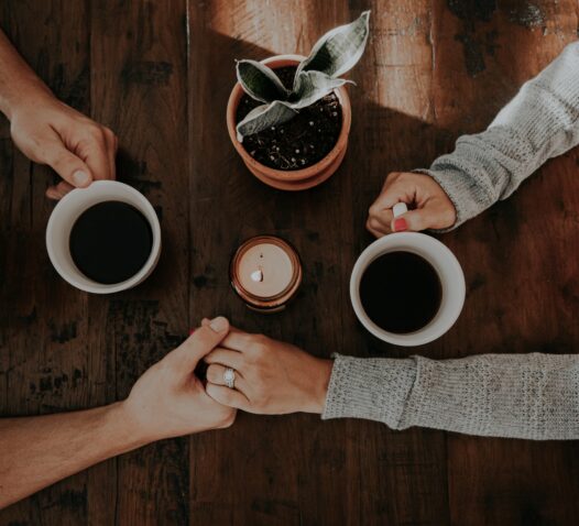 Två personer håller varandra i handen över ett bord med två kaffekoppar och en liten växt.